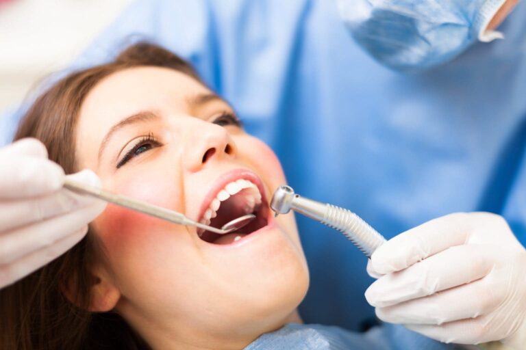 Dentist wearing a blue surgical gown and face mask, using dental tools on a patient lying back in a dental chair with a dental bib on.