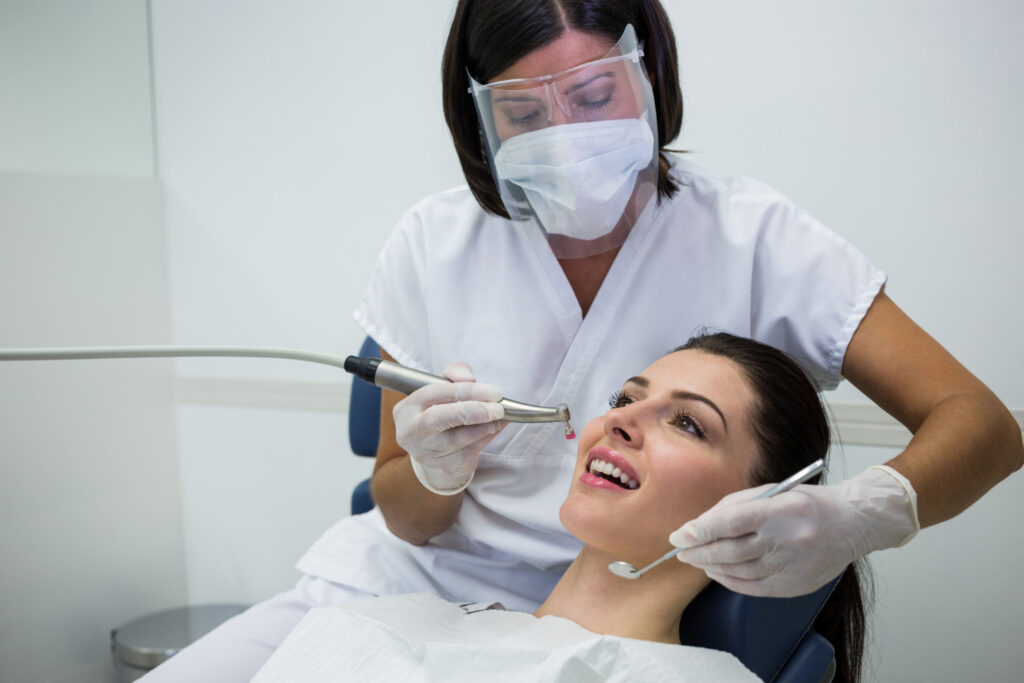 Dental professional in white scrubs using dental instruments on a patient reclining in a dental chair, with a protective bib.