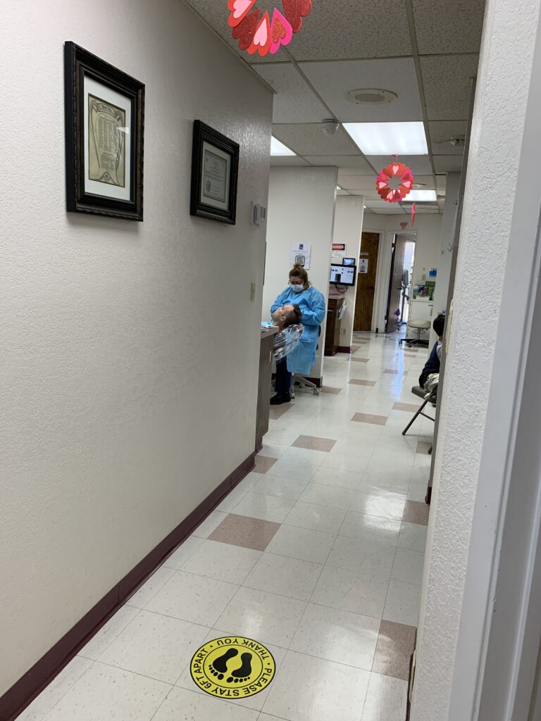Clean, modern hallway with tiled floor and heart-shaped ceiling decorations at Art Family Dental clinic. A person in a protective gown and mask attending to a patient in a dental chair, emphasizing safety and care during dental procedures.