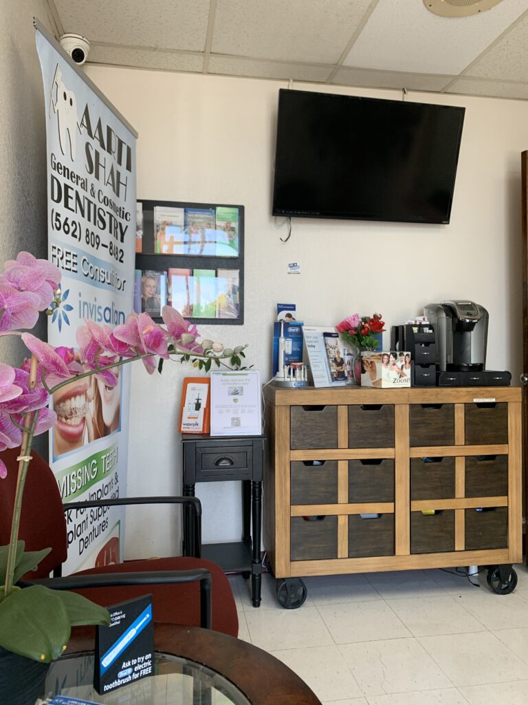Waiting area in a dental office with a banner advertising Aarti Shah General & Cosmetic Dentistry, a Keurig coffee machine, brochures, a wall-mounted TV, and decorative items like an orchid plant and framed displays.