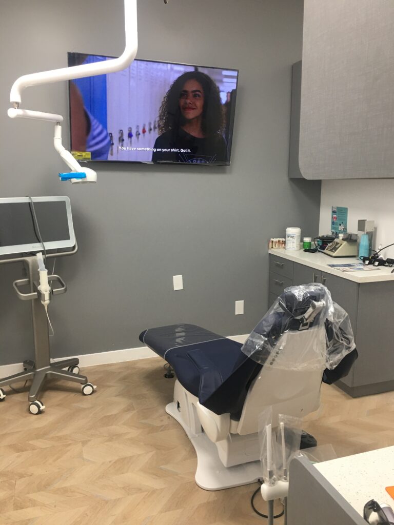Modern dental office with a plastic-covered dental chair, mounted TV displaying a show, dental light, various equipment, and dental supplies on cabinets and countertops. The floor has a wood-like pattern, and the walls are painted gray.