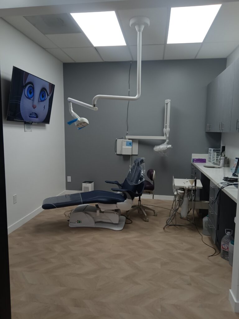 Modern dental room with a dental chair, equipment, and a TV screen displaying an animated character. The room is well-lit, with white and gray walls, light wood flooring, and various dental supplies and tools.